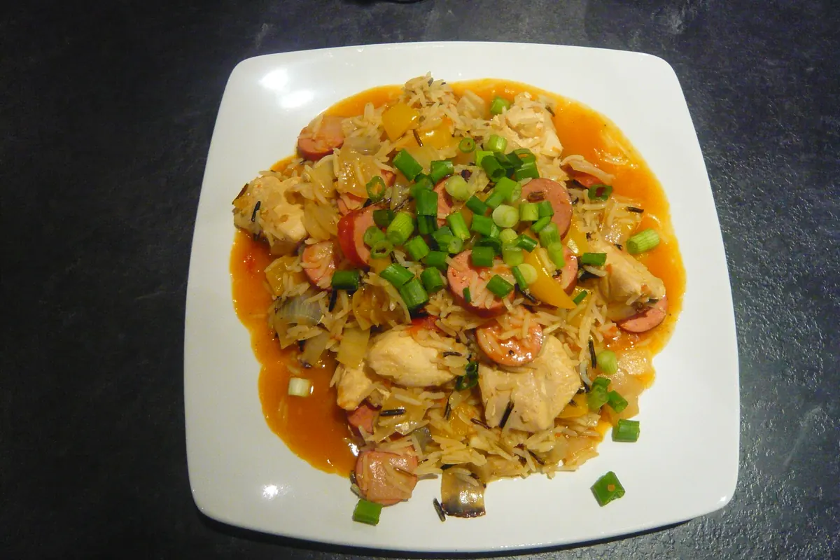 A white square plate of chicken and sausage jambalaya in a tomato sauce, topped with chopped spring onion