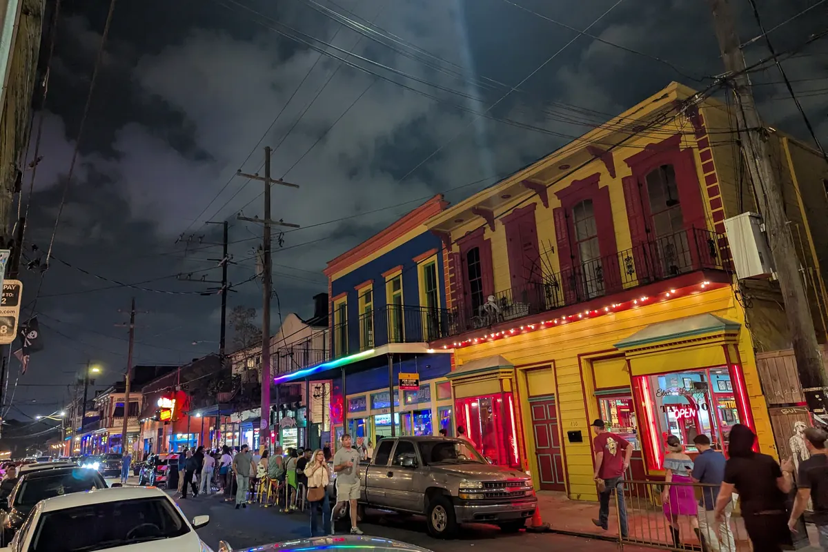 Frenchmen Street at night, with brightly painted red, blue and yellow buildings, neon signs and a queue of people on the pavement under a dark cloudy sky